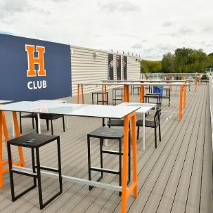 The open sky deck features an H-Club panel  on the wall as well as high top tables and stool for people to use while watching a game.